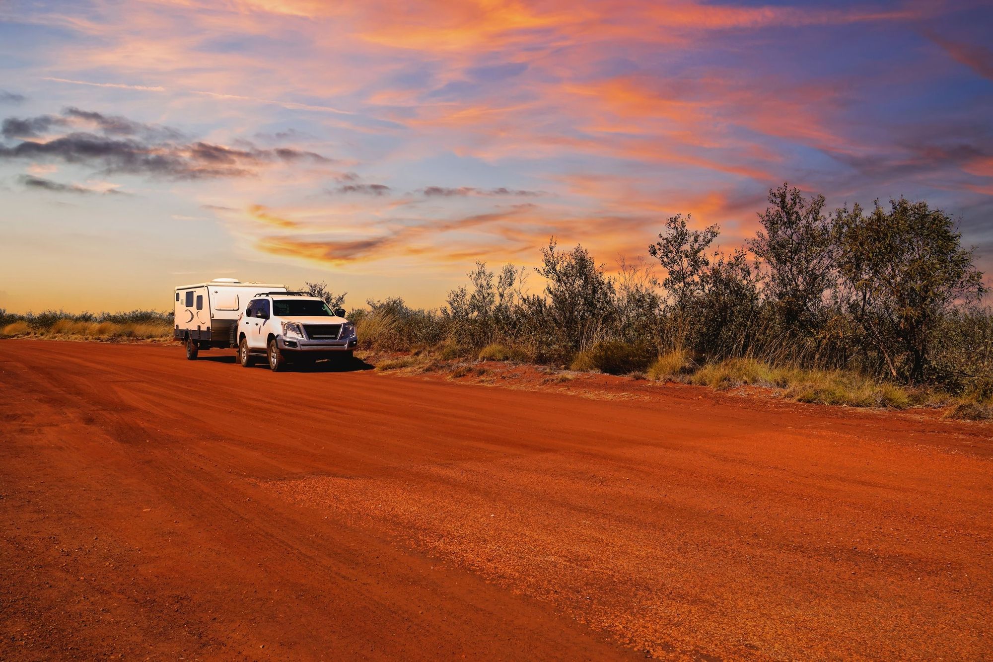 White SUV towing a caravan on a red dirt road at sunset